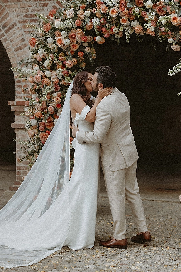 Wedding kiss portrait of bride and groom kissing beneath a rose floral arch, her long veil flowing by a brick archway on a stone walkway