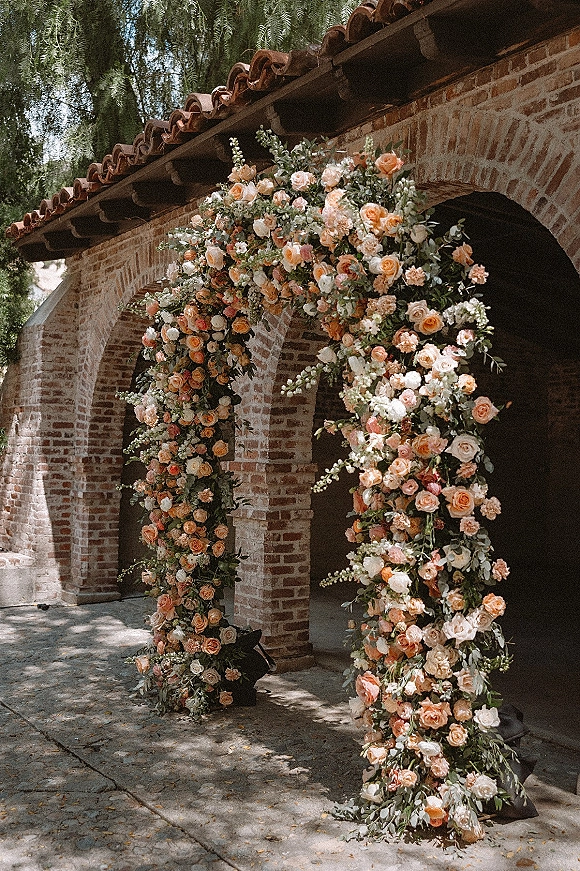 Floral wedding arch with asymmetrical blush and peach roses, white blooms, and greenery set against brick arches in dappled sunlight