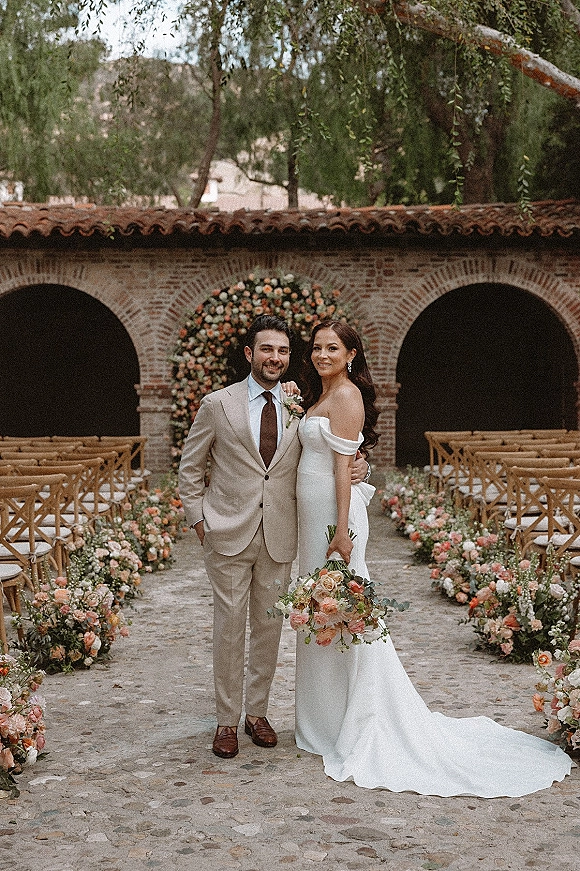 Couple portrait of bride holding bouquet beside groom in a beige suit, standing on a stone walkway under brick arches in a courtyard