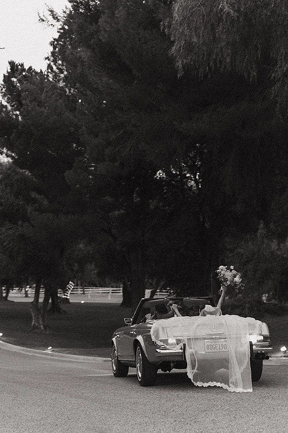 Wedding getaway car with a bride and groom in a convertible, veil and ribbon trailing, bouquet and flowers on a tree-lined road