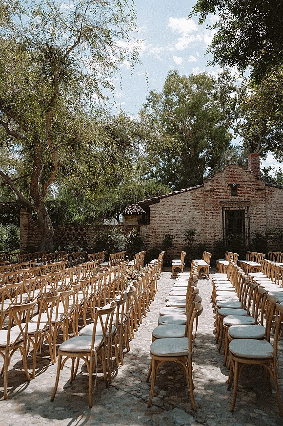 Outdoor ceremony setup with wood chairs and cushions flanking a double aisle with floral clusters in a sunny brick courtyard