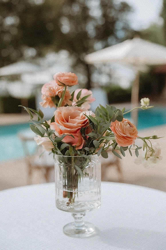Wedding centerpiece of peach flowers and greenery in a clear glass vase on a white tablecloth, set on a poolside patio with umbrellas