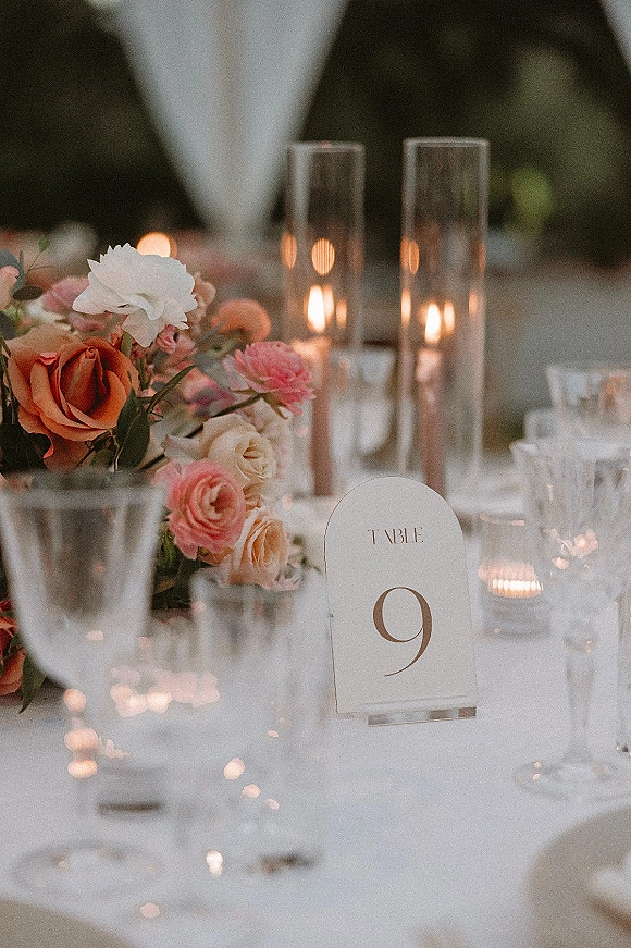Reception tablescape with wedding table centerpiece of roses, taper and hurricane candles, champagne flutes on white linens under bistro lights