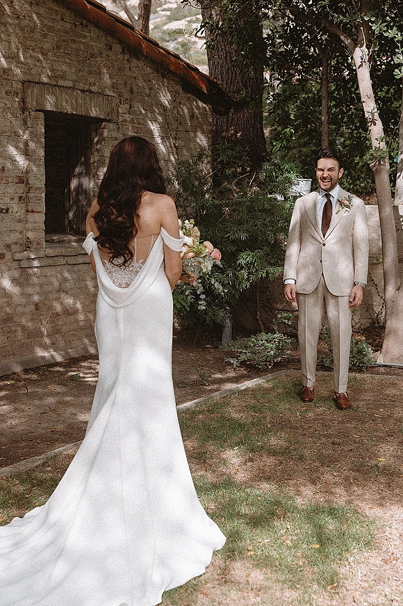 Wedding first look as bride in an off the shoulder gown with long train walks to groom in beige suit by rustic brick building in dappled light