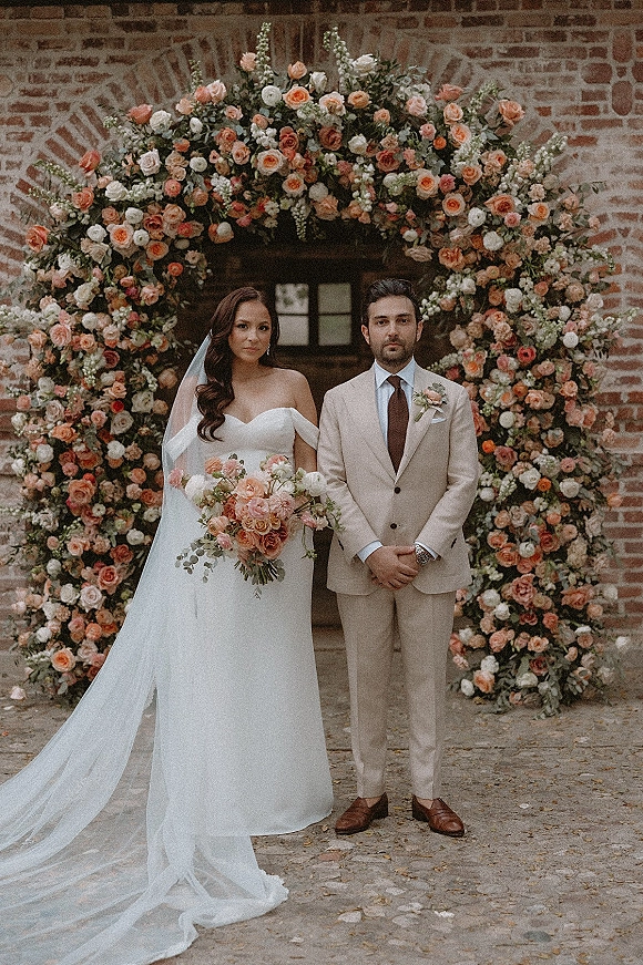 Couple portrait of bride and groom portrait under a rose floral arch, bride in strapless gown holding bouquet before a brick wall door