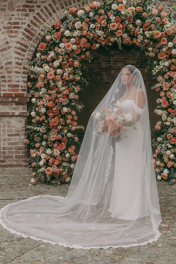 Bridal portrait of a bride with veil in a strapless wedding dress, holding a bouquet beneath a rose floral arch in a brick courtyard