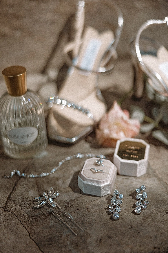 Bridal accessories wedding detail flat lay with heels, perfume bottle, rings in boxes, crystal earrings, hair pins and petals on stone surface