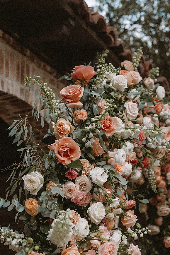 Wedding floral arch with rose wedding arch blooms in peach and white, eucalyptus greenery, set against a brick wall with roof tiles and trees