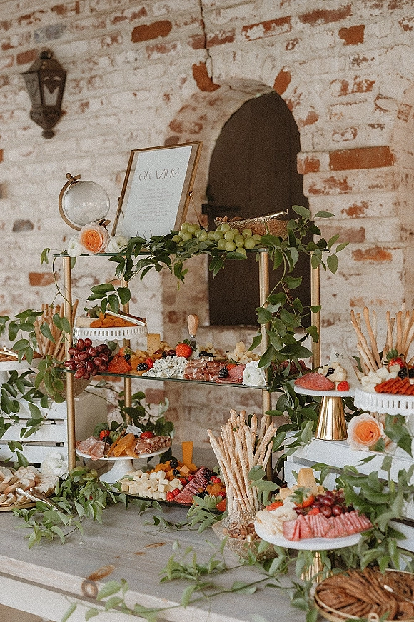 Wedding grazing table with charcuterie table wedding spreads of cheese, fruit, crackers, and greenery accents against a whitewashed brick wall