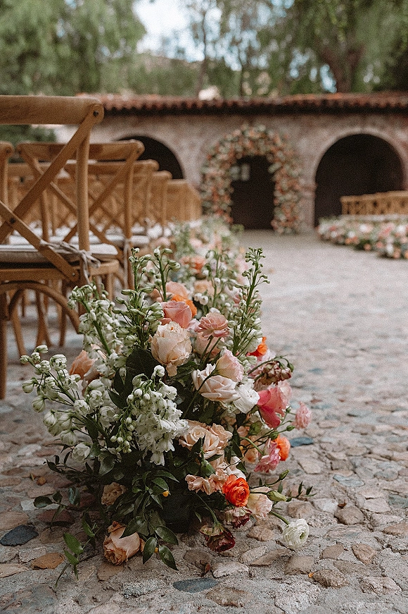 Ceremony aisle flowers in low ground clusters lining a cobblestone path, with a floral arch and wooden crossback chairs in a stone courtyard