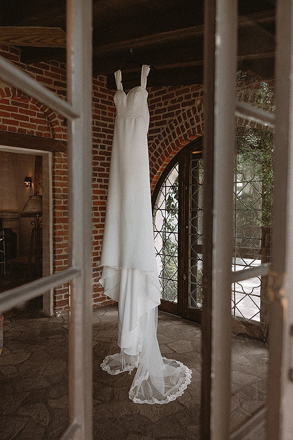 Wedding dress hanging on a hanger, strapless bridal gown with lace hem and tulle train framed by arched leaded glass doors and brick wall