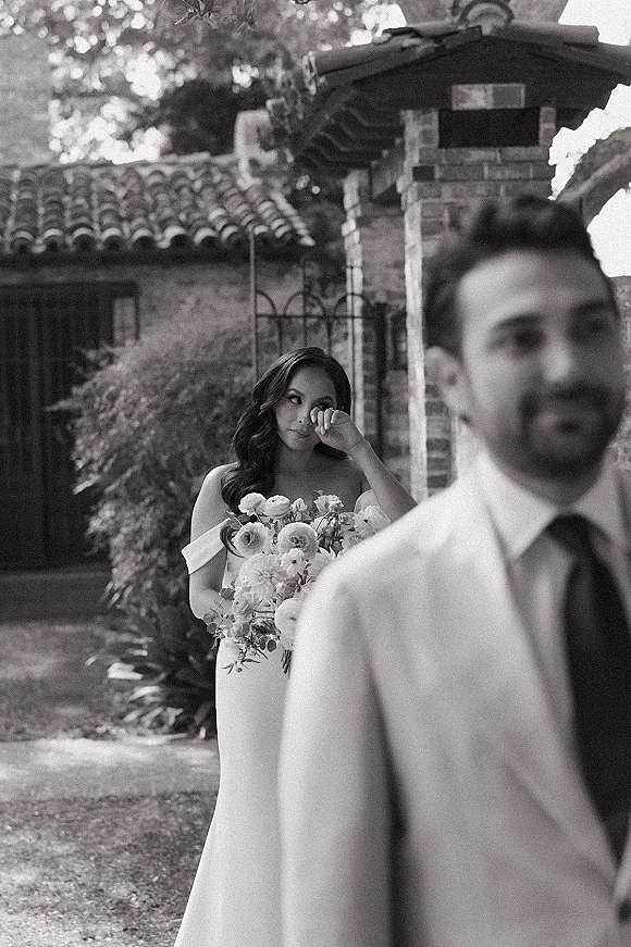 Wedding first look as bride behind groom wipes tears holding a bouquet in an off-the-shoulder dress by a stone gate garden courtyard