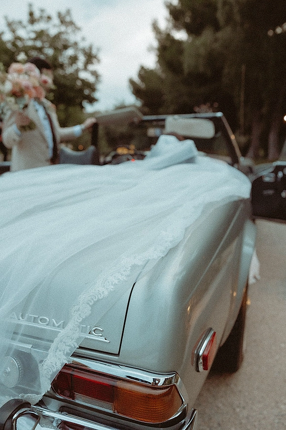 Wedding getaway car, a vintage convertible with the bride’s lace dress train and bouquet draped over the side on a tree-lined road