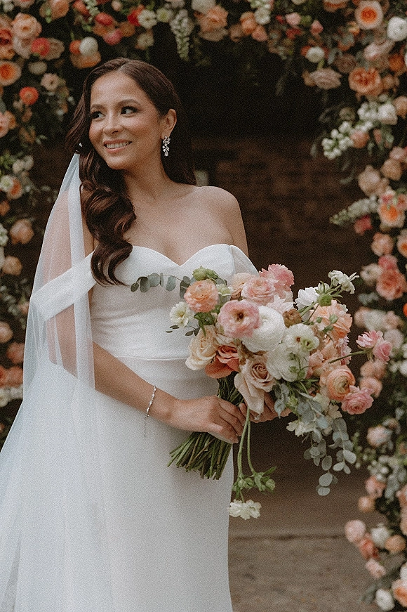 Bridal portrait of a bride holding bouquet in a strapless wedding dress, framed by a floral arch near a brick doorway and flower wall backdrop