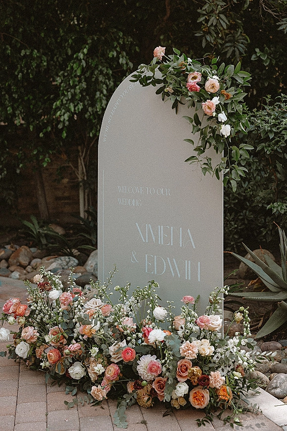 Wedding welcome sign in an arched design with peach roses, ranunculus and eucalyptus, set along a paver garden path by a stone wall