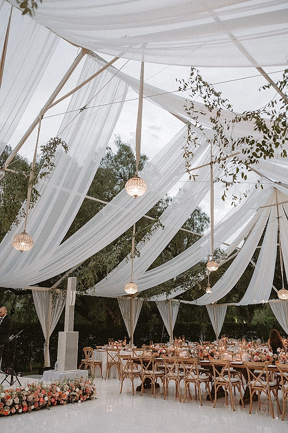 Reception decor with sheer draping canopy and hanging lanterns over long banquet tables with greenery garlands, candles, and floral centerpieces in a garden setting
