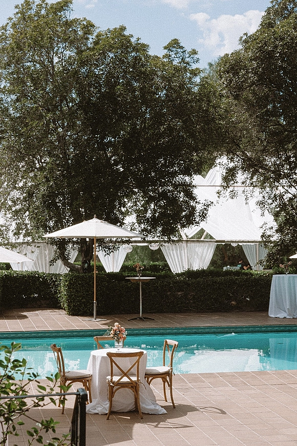 Poolside reception setup with a round table, white tablecloth and floral centerpiece under a patio umbrella beside the swimming pool and tent
