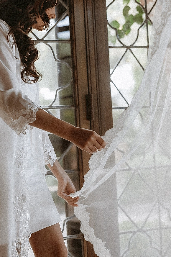 Bride getting ready in a white lace robe, holding her bridal veil detail by a lead glass window in soft natural light