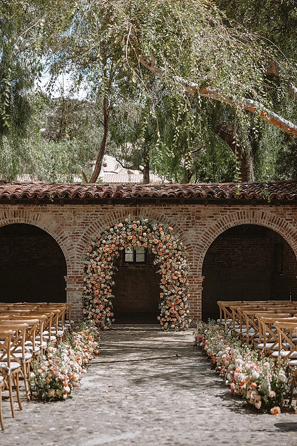 Ceremony setup for an outdoor wedding ceremony with a round floral arch, flower-lined aisle, and wooden chairs in a brick-arched courtyard
