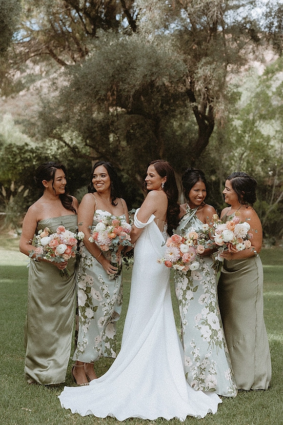 Bridesmaid group portrait with bride with bridesmaids holding blush bouquets, laughing in mismatched satin and floral dresses on a garden lawn