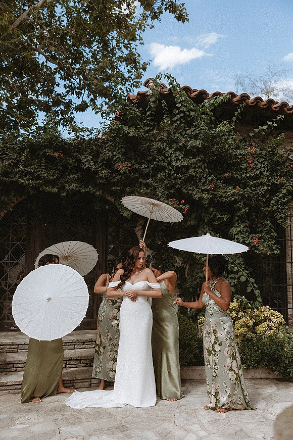 Bride with bridesmaids holding white parasol as they gather on stone steps by an ivy-covered wall in a sunny courtyard under blue sky