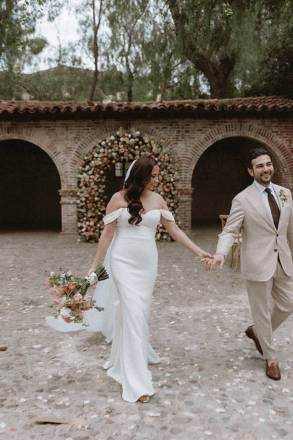 Couple portrait of bride and groom holding hands beneath a wedding floral arch in a brick-arch courtyard, framed by scattered petals