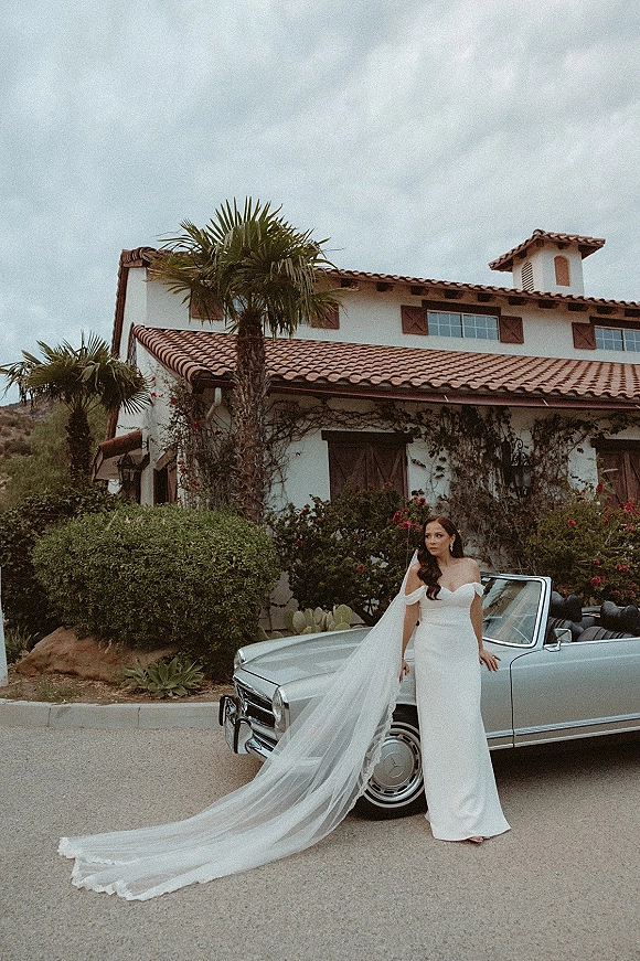 Bridal portrait of a bride in a strapless wedding dress with a long cathedral veil, leaning on a classic convertible by a villa driveway