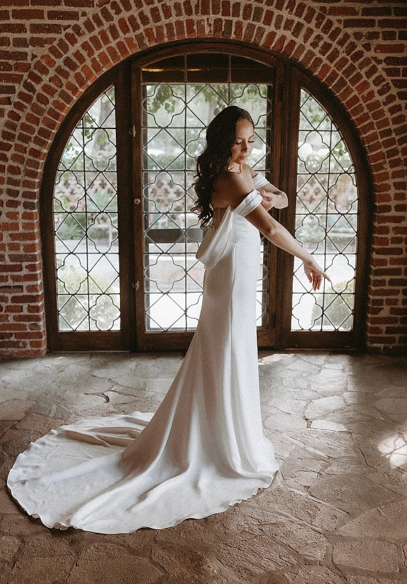 Bridal portrait of a bride in an off the shoulder wedding dress with a long train, standing by an arched window against a brick wall