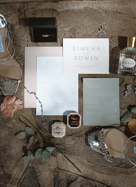 Invitation suite wedding flat lay with wax-sealed envelope, rings in velvet box, perfume, jewelry, shoes, and rose on stone surface