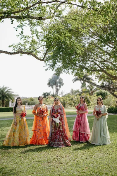 Bride with bridesmaids in colorful lehengas, bride in red lehenga and veil with jewelry, holding red bouquets on a garden lawn under trees