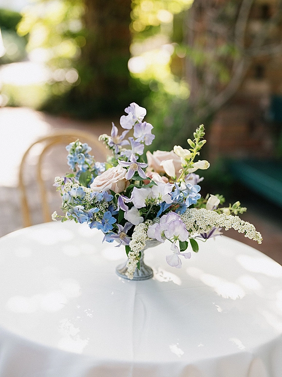 Wedding centerpiece with blush roses and blue flowers in a small vase on a round white table in a sunlit garden setting