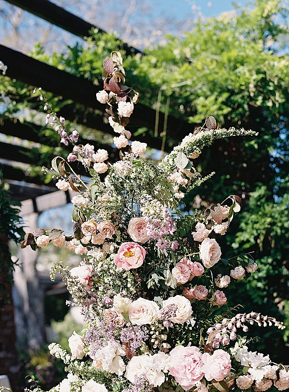 Wedding floral arrangement ceremony floral arrangement with pastel roses and peonies, lush greenery spray beneath pergola beams and blue sky