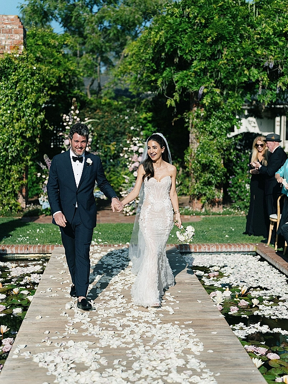Wedding recessional as bride and groom walking hand in hand down a rose-petal wooden walkway beside a lily pad pond, guests seated nearby