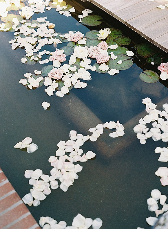 Floating flower decor with floating rose petals and blush roses drifting beside lily pads on a pond near a wooden deck edge