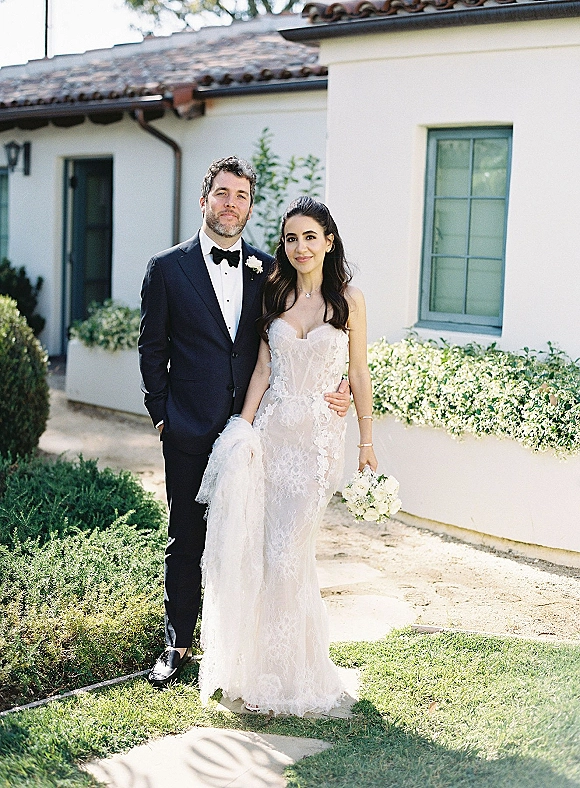 Couple portrait of bride and groom portrait with strapless lace gown and tuxedo, holding white bouquet outside stucco villa with tiled roof
