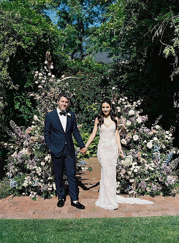 Couple portrait of bride and groom holding hands beneath a floral arch of roses, on a brick pathway in a lush garden under blue sky