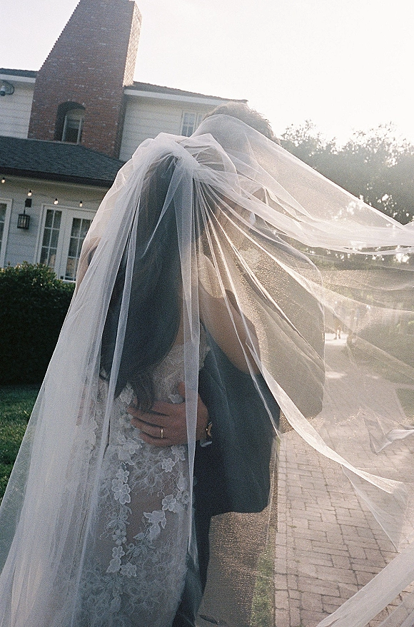 Wedding couple portrait with bride and groom embrace under a long veil, backlit by sunlight near a house with string lights and lawn walkway