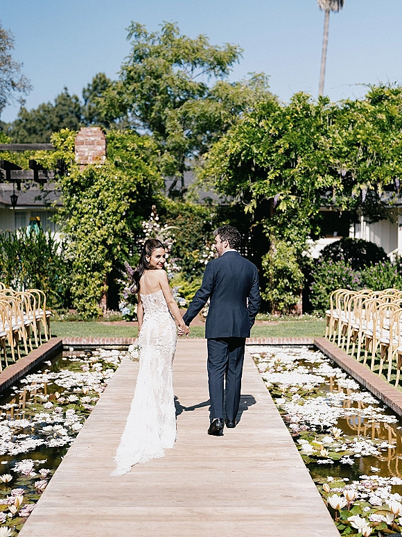 Couple portrait of newlyweds holding hands, walking away on a wooden walkway over a lily pond with ceremony chairs and garden greenery under blue sky