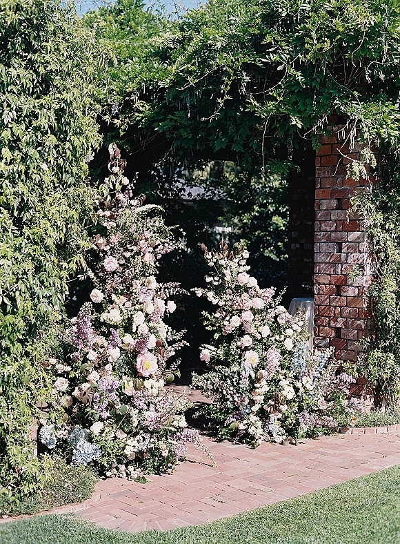 Ceremony floral arch with lush rose and hydrangea arrangements, greenery garland and vines framing a brick pillar garden walkway