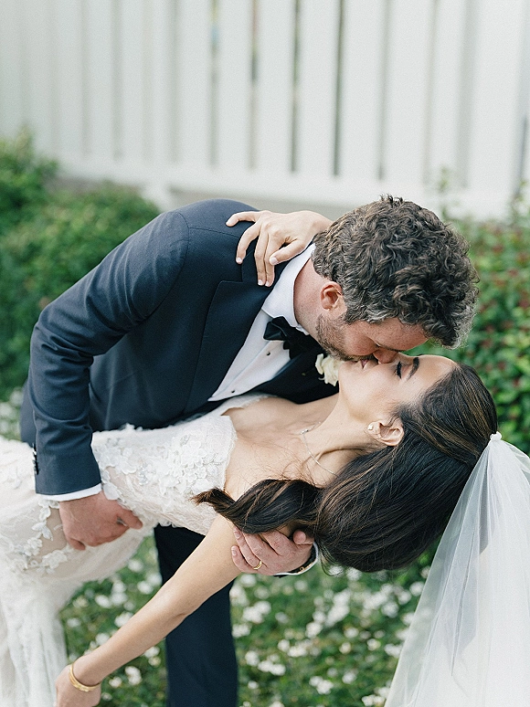 Wedding kiss portrait of groom dipping bride in veil and lace dress as they kiss, framed by garden greenery and a white fence backdrop