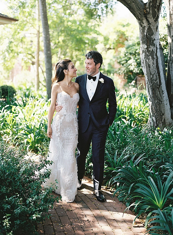 Couple portrait of bride and groom walking on a brick path, gazing at each other amid garden greenery, bride in strapless lace dress