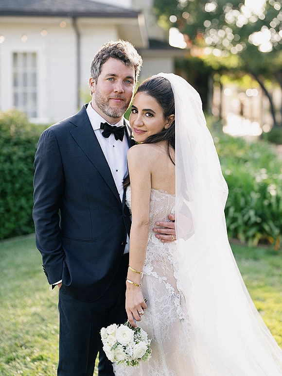 Couple portrait of bride and groom portrait outdoors, bride in lace dress and veil with white rose bouquet beside tuxedoed groom on lawn
