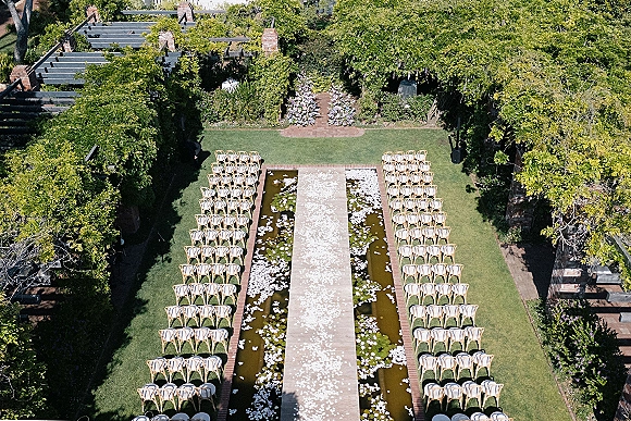 Ceremony setup with outdoor ceremony seating, wood chairs lining an aisle runner with flower petals beside a garden pergola and lawn