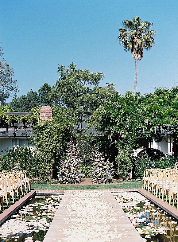 Ceremony aisle decor with petal-strewn walkway over a reflecting pool, lined by wood chairs and pastel florals beneath an arch in a garden courtyard