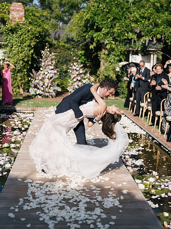 Wedding kiss portrait of groom dipping the bride on a petal-covered wooden aisle runner by a lily pond, veil flowing as guests cheer