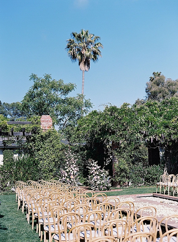 Ceremony setup for an outdoor wedding ceremony with wood chairs, white cushions, and a flower petal aisle leading to a pergola on a garden lawn