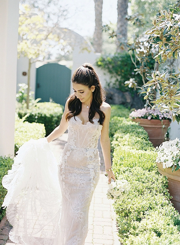 Bridal portrait of a bride holding bouquet in a strapless lace wedding dress with long train, looking down on a garden walkway with hedges