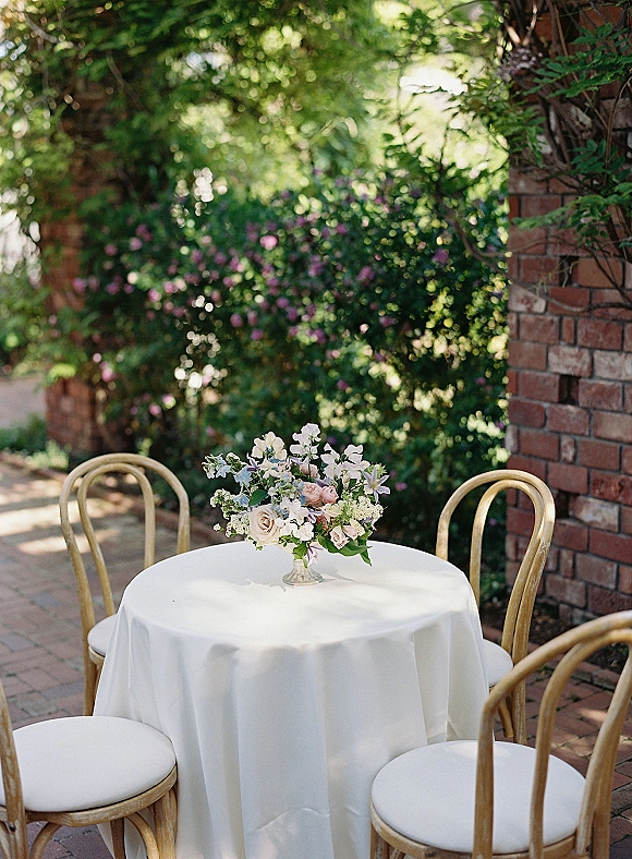 Reception tablescape with a round wedding table setup, white linen, pedestal vase floral centerpiece, and wooden chairs on a brick patio garden