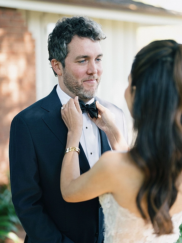 First look moment as bride fixes groom’s bow tie, her engagement ring visible beside his black tuxedo by a brick wall with greenery