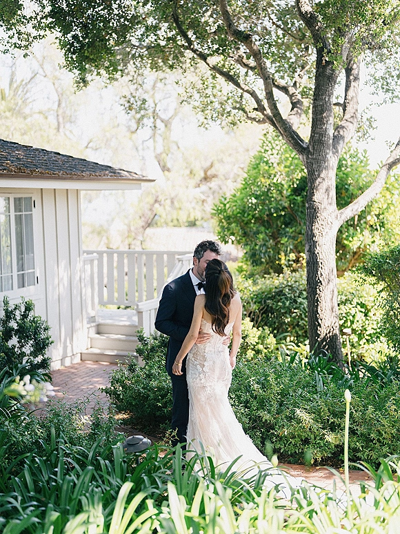 Wedding couple portrait of bride and groom embrace on a brick garden path by a white cottage, bride in a lace wedding dress and groom in tuxedo bow tie